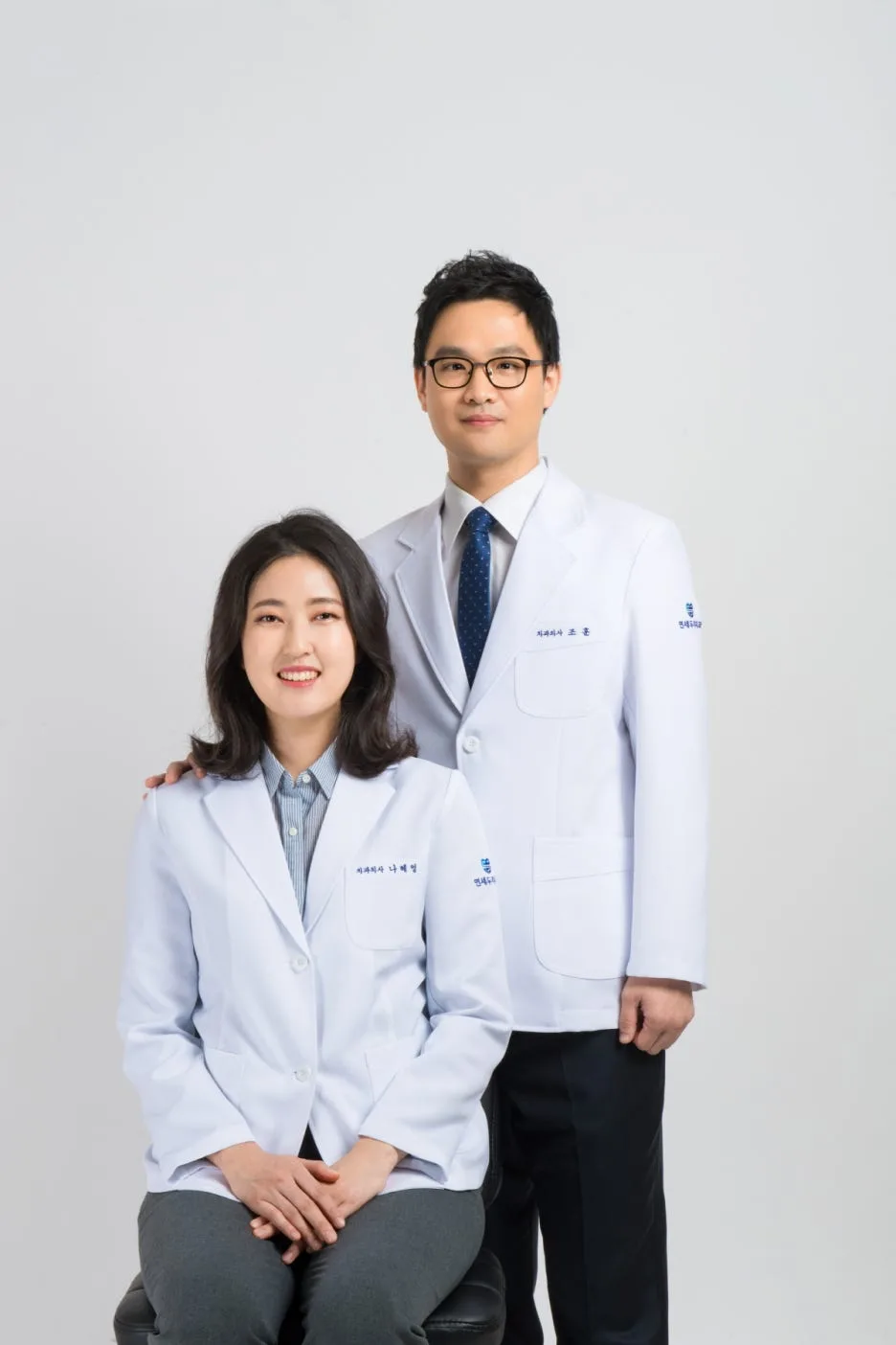 임플란트 뼈이식 - 치과 의사 두 명이 흰색 의사 가운을 입고 카메라를 보고 서 있다.